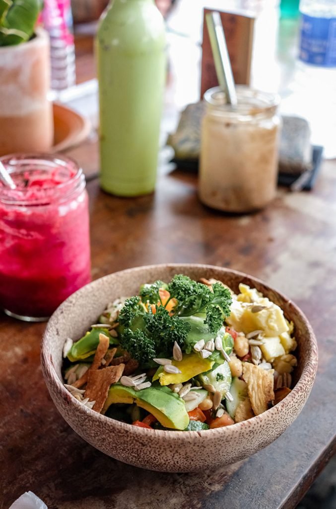 Close-up of a vibrant and healthy vegan meal in a ceramic bowl, featuring broccoli, avocado slices, and a variety of seeds, with drinks in the background. Smoothies are one example of how Sri Lanka is not expensive.