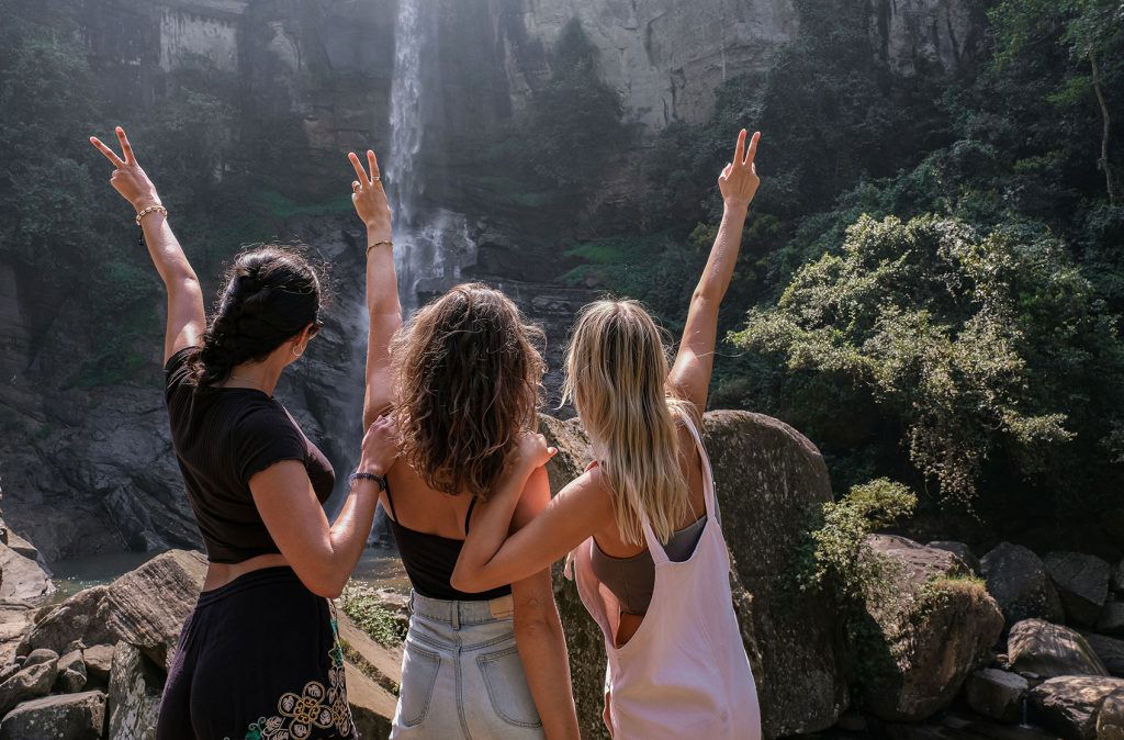 Three women celebrating at a waterfall, raising their hands in the air with peace signs, surrounded by lush greenery